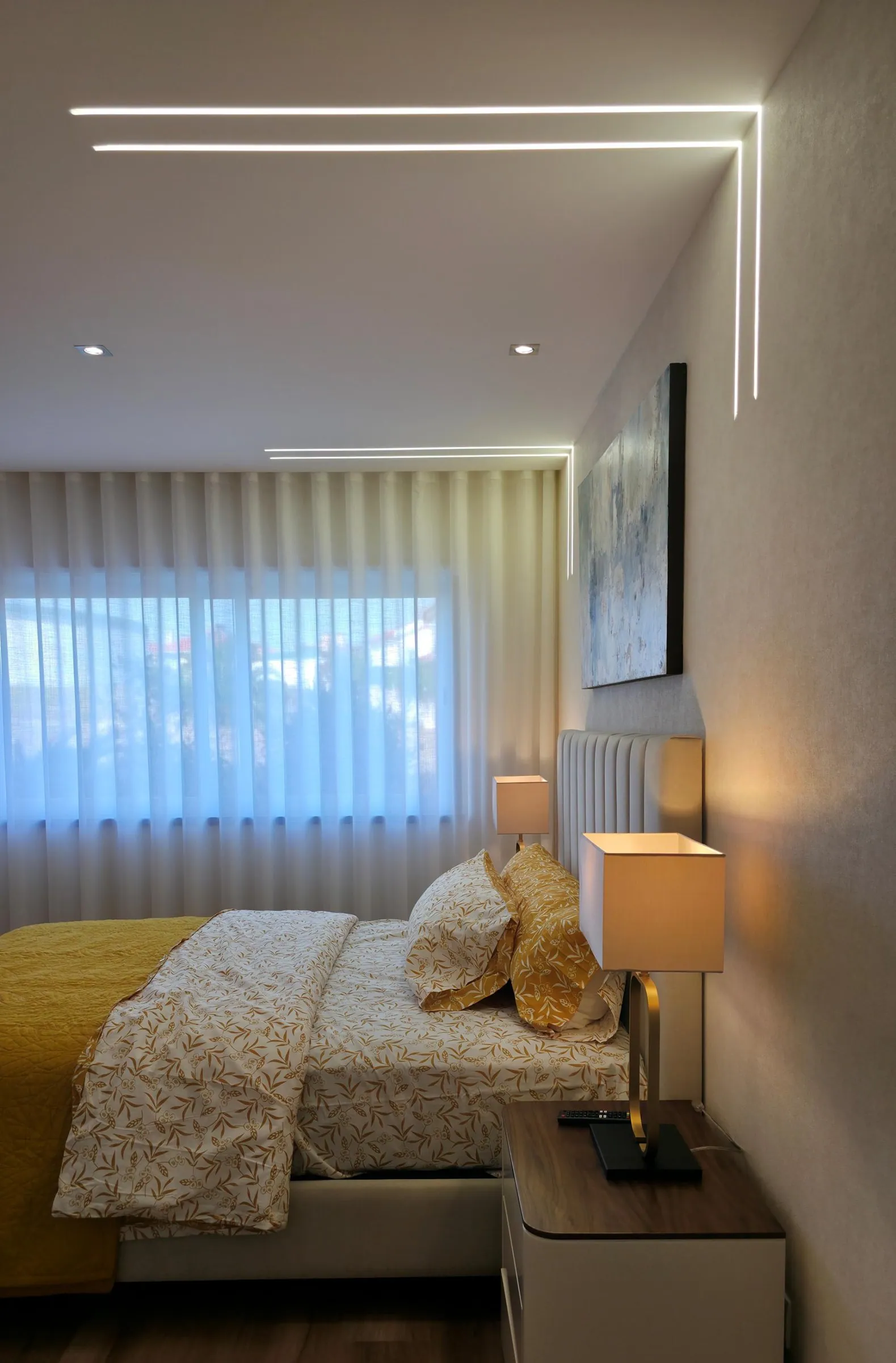 Quarto com colcha mostarda, candeeiros de mesa e cortinados claros.